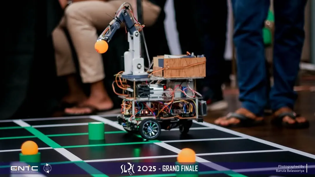 The robot on the SLRC arena floor, its arm holding an orange ball above a grid of black and green lines.